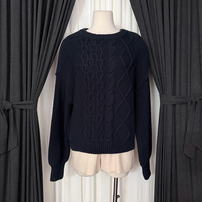 May include: A navy blue cable-knit sweater with a crew neck. The sweater features a textured pattern on the front and sleeves. The sleeves are long and the hemline is ribbed. The sweater is displayed on a mannequin.