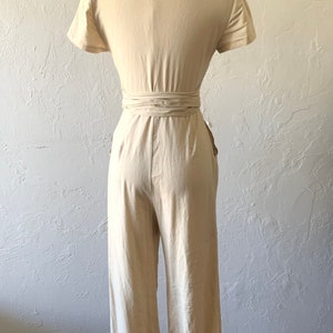 May include: A light beige linen jumpsuit with a wrap tie at the waist and short sleeves. The jumpsuit has pockets on the sides.