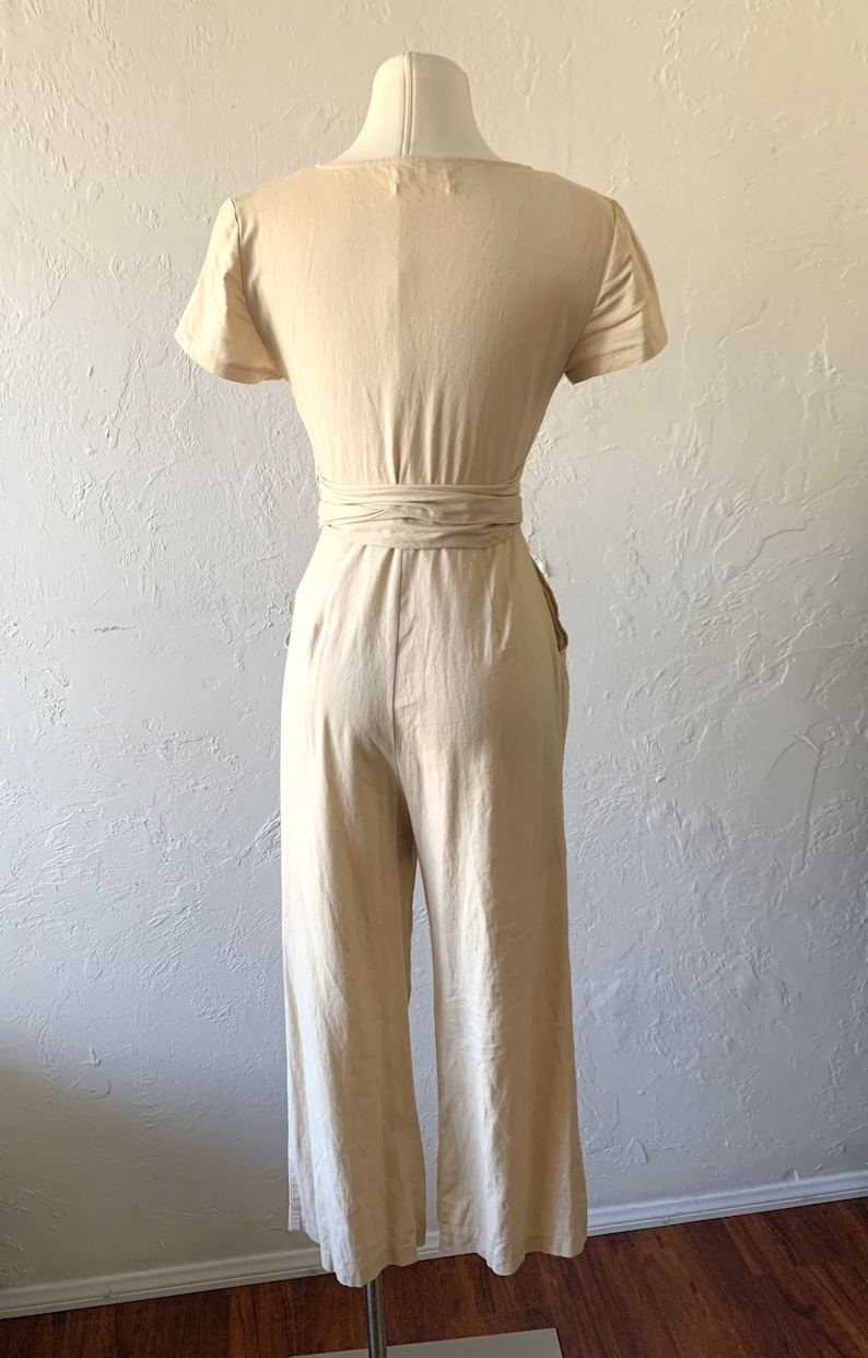 May include: A light beige linen jumpsuit with a wrap tie at the waist and short sleeves. The jumpsuit has pockets on the sides.
