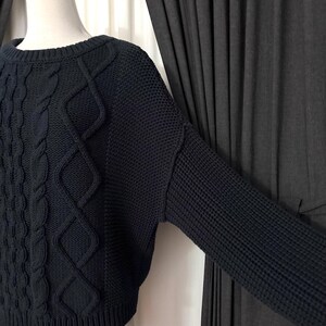 May include: A navy blue sweater with a crew neck and long sleeves. The front features a cable knit design with diamond-shaped patterns. The sleeves have a waffle knit texture. The sweater is displayed on a mannequin.