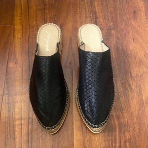 May include: A pair of black leather slip-on shoes with a snakeskin pattern. The shoes have a pointed toe and a woven jute sole. The brand name "Free People" is visible on the inside of the shoes.