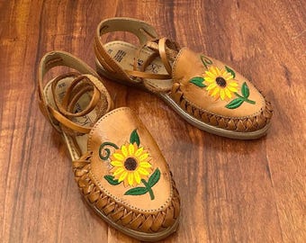 May include: A pair of brown leather sandals with sunflower embroidery. The sandals have a woven strap and a lace-up closure.