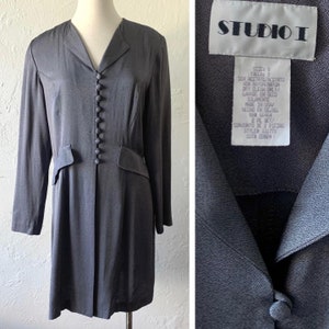 May include: A long-sleeved, gray dress with a button-up front and a v-neck collar. The dress has a Studio I label with care instructions and size information. The label reads: "Size: 8 Talla: S Sex Acetate/Acetato 4SX Rayon/Raton Dry Clean Only/ Lavado en Seco Solamente Made in USA/ Hecho en EE.UU. RN# 66464 2 PC SET/ Conjunto de 2 Piezas Style# 33177x Cut# 22824"
