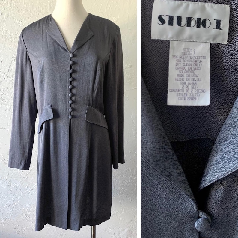 May include: A long-sleeved, gray dress with a button-up front and a v-neck collar. The dress has a Studio I label with care instructions and size information. The label reads: "Size: 8 Talla: S Sex Acetate/Acetato 4SX Rayon/Raton Dry Clean Only/ Lavado en Seco Solamente Made in USA/ Hecho en EE.UU. RN# 66464 2 PC SET/ Conjunto de 2 Piezas Style# 33177x Cut# 22824"