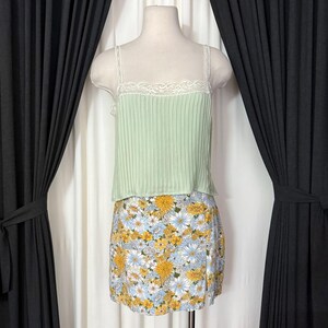 May include: A mint green camisole with a pleated design and lace trim, paired with a floral print mini skirt. The skirt features a pattern of yellow and blue flowers on a white background. The outfit is displayed on a mannequin.
