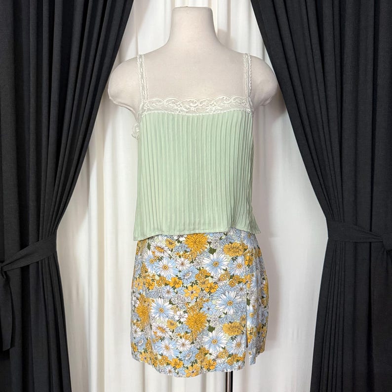 May include: A mint green camisole with a pleated design and lace trim, paired with a floral print mini skirt. The skirt features a pattern of yellow and blue flowers on a white background. The outfit is displayed on a mannequin.