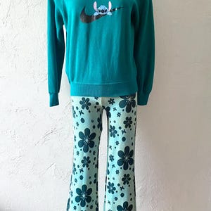 K&ouml;nnte beinhalten: T&uuml;rkisblauer Pullover mit einem schwarzen Nike-Swoosh und einem blauen Stitch-Charakter im Swoosh. Der Pullover wird mit hellblauen Hosen mit einem dunkelblauen Blumenmuster kombiniert.
