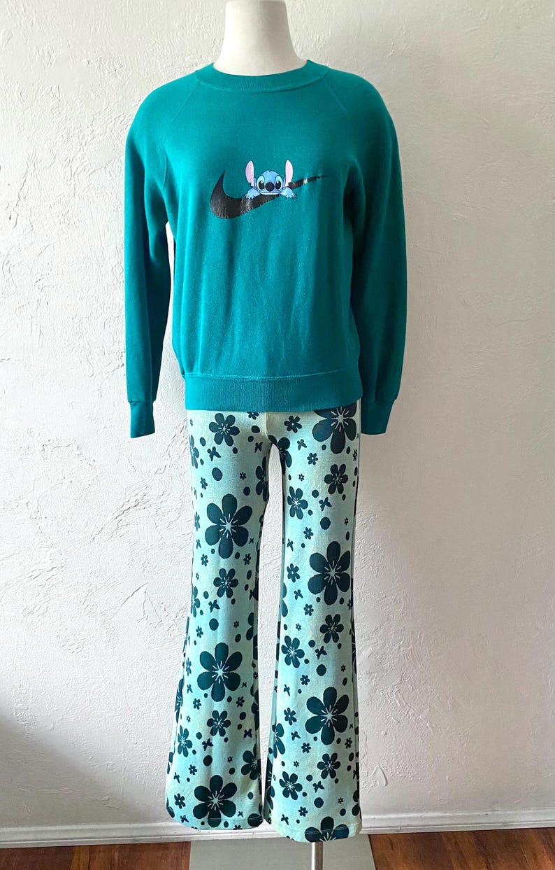 K&ouml;nnte beinhalten: T&uuml;rkisblauer Pullover mit einem schwarzen Nike-Swoosh und einem blauen Stitch-Charakter im Swoosh. Der Pullover wird mit hellblauen Hosen mit einem dunkelblauen Blumenmuster kombiniert.