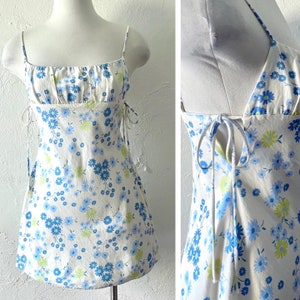 May include: A white mini dress with a blue and green floral print. The dress has thin straps and a tie at the back.