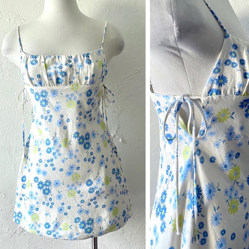 May include: A white mini dress with a blue and green floral print. The dress has thin straps and a tie at the back.