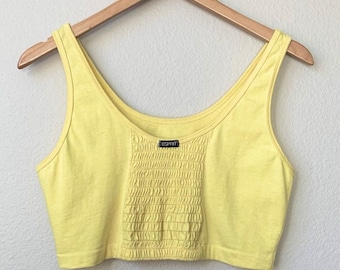 Pode incluir: Um top amarelo de alças com um detalhe franzido na frente. O top tem um decote redondo e é feito de um tecido macio e elástico. A etiqueta da marca diz "Esprit".