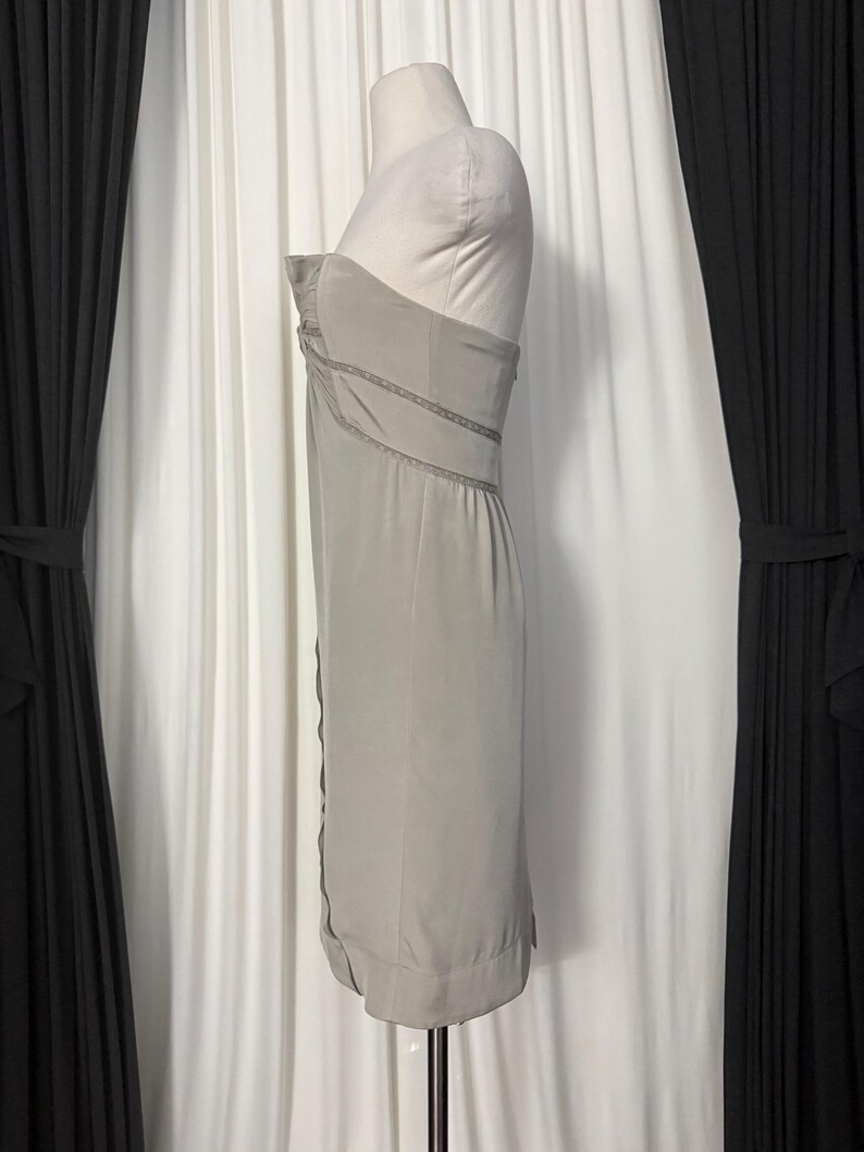 May include: A strapless, knee-length dress in a muted sage green color. The garment features a fitted bodice with subtle vertical pleating and delicate lace trim detailing along the seams, creating a structured silhouette on a dress form.
