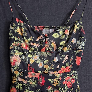May include: Black floral print dress with spaghetti straps. The dress features a vibrant pattern of red, orange, yellow, and green flowers on a black background. It has a fitted bodice with small buttons and a gathered neckline.