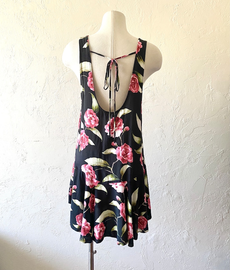 May include: A black sleeveless dress with a floral print featuring pink roses and green leaves. The dress has a tie back and a ruffled hem.