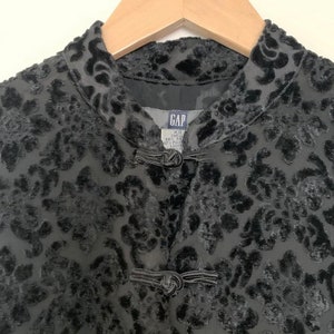 May include: Black velvet jacket with a floral pattern and a mandarin collar. The jacket has two buttons on the front and a label that reads "GAP XS SHELL 60% POLY 40% RAYON LINING 100% POLY".