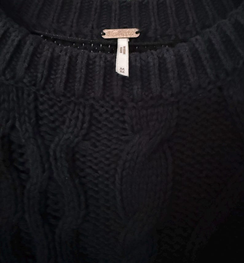 May include: Close-up of a black knit sweater with a cable knit design. The sweater has a ribbed neckline and a small metal tag with the brand name. The tag is centered at the back of the neck. The sweater is a size small.