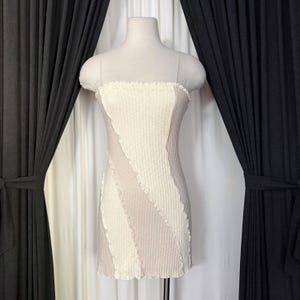 May include: A cream-colored, ribbed mini dress with a unique design. The dress features a strapless bodice and thin straps. The dress has a fitted silhouette with diagonal panels of varying shades of cream and beige. The hemline has a scalloped edge.