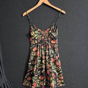 May include: A black floral print dress with spaghetti straps hangs on a wooden hanger. The dress features a vibrant pattern of red, yellow, and green flowers. The fabric appears to be lightweight, with a fitted bodice and a flared skirt.