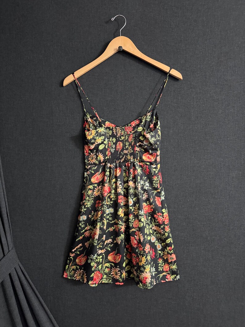 May include: A black floral print dress with spaghetti straps hangs on a wooden hanger. The dress features a vibrant pattern of red, yellow, and green flowers. The fabric appears to be lightweight, with a fitted bodice and a flared skirt.