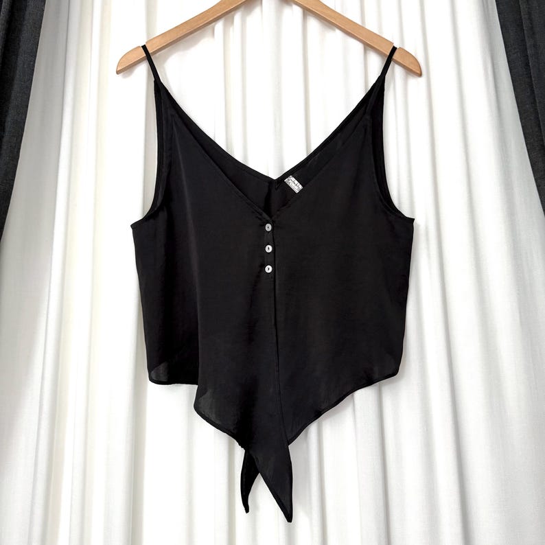 May include: Black camisole top with a V-neckline and spaghetti straps. The top features three small buttons down the front and a tie detail at the bottom. The garment is hanging on a wooden hanger against a white backdrop.