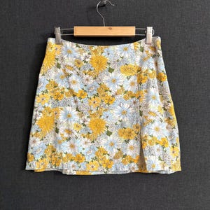 May include: A floral print mini skirt featuring a pattern of yellow and white daisies and blue flowers on a white background. The skirt is hanging on a wooden hanger against a gray background.