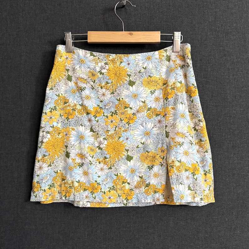May include: A floral print mini skirt featuring a pattern of yellow and white daisies and blue flowers on a white background. The skirt is hanging on a wooden hanger against a gray background.