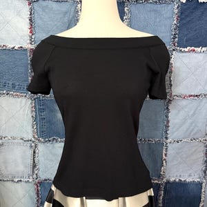 May include: A black off-the-shoulder top with short sleeves. The top is displayed on a mannequin, paired with a black and white striped skirt. The background is a patchwork of denim squares.
