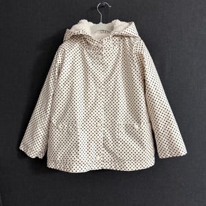 May include: Cream-coloured jacket with black polka dots, a hood, and a soft, white, fuzzy lining. The jacket has a button-up front, two pockets, and is hanging on a metal hanger. A stylish outerwear piece.