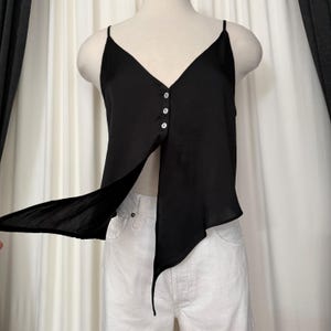 May include: Black camisole top with spaghetti straps and a V-neckline. The top features three buttons down the center and an asymmetrical design with a flowing, open front. The garment is displayed on a mannequin.