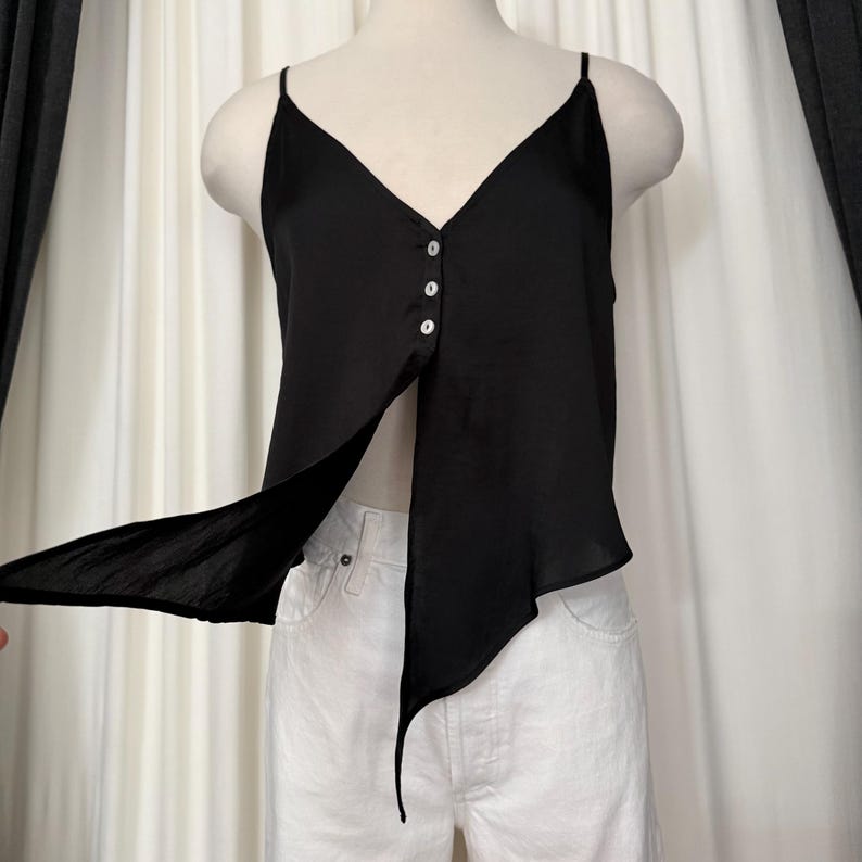 May include: Black camisole top with spaghetti straps and a V-neckline. The top features three buttons down the center and an asymmetrical design with a flowing, open front. The garment is displayed on a mannequin.