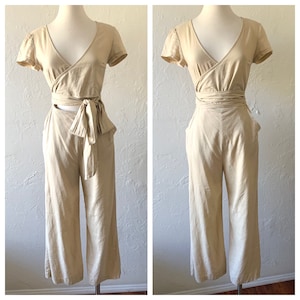 May include: A beige linen jumpsuit with a wrap top and wide-leg pants. The top has a tie at the waist and a cutout at the back. The pants have pockets on the sides.