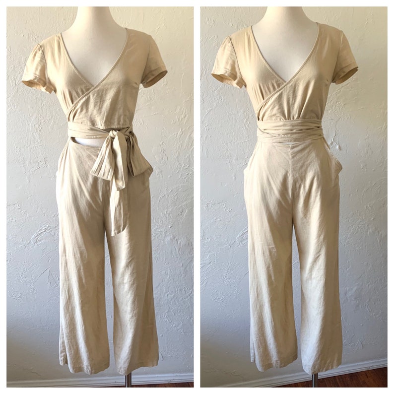 May include: A beige linen jumpsuit with a wrap top and wide-leg pants. The top has a tie at the waist and a cutout at the back. The pants have pockets on the sides.