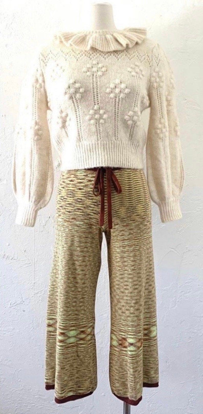 May include: A white knitted sweater with a ruffled collar and a brown and beige knitted wide-leg pant.