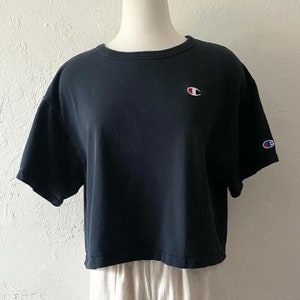 Puede incluir: Camiseta negra de manga corta y corte corto con un pequeño logotipo de Champion en la parte delantera y un logotipo más grande en la manga derecha.