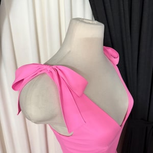 May include: Bright pink dress with a V-neckline and large bow details on the shoulders. The dress is displayed on a mannequin, showcasing the design and fit. The fabric appears smooth and the color is vibrant.