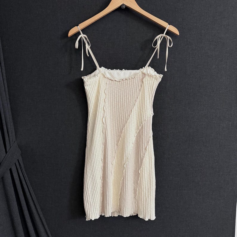 May include: Cream-colored ribbed knit dress with tie straps. The dress features a ruffled neckline and decorative stitching. The dress is hanging on a wooden hanger against a dark background.