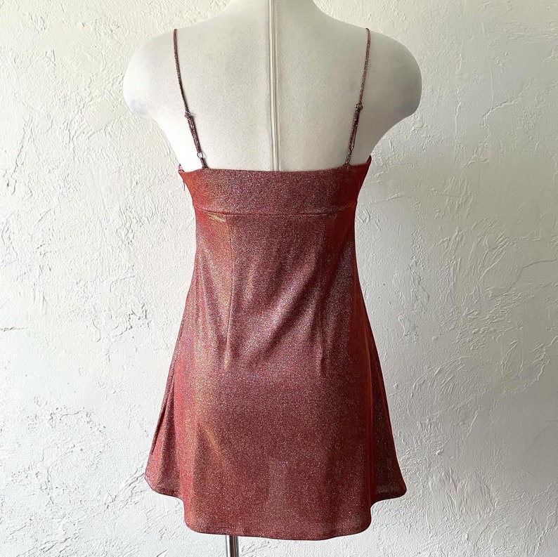 Puede incluir: Un minivestido brillante de color marr&oacute;n rojizo con finos tirantes espagueti. El vestido tiene un escote cuadrado y una falda ligeramente acampanada. La tela tiene un sutil efecto brillante, que capta la luz.