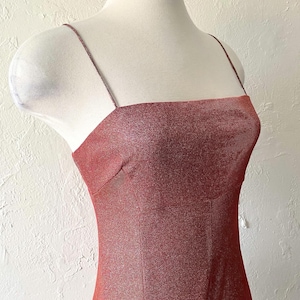 Puede incluir: Vestido rojo-marr&oacute;n brillante con finos tirantes espagueti y escote cuadrado. El vestido se exhibe en un maniqu&iacute;, mostrando su silueta ajustada. La tela tiene una apariencia sutil y texturizada.