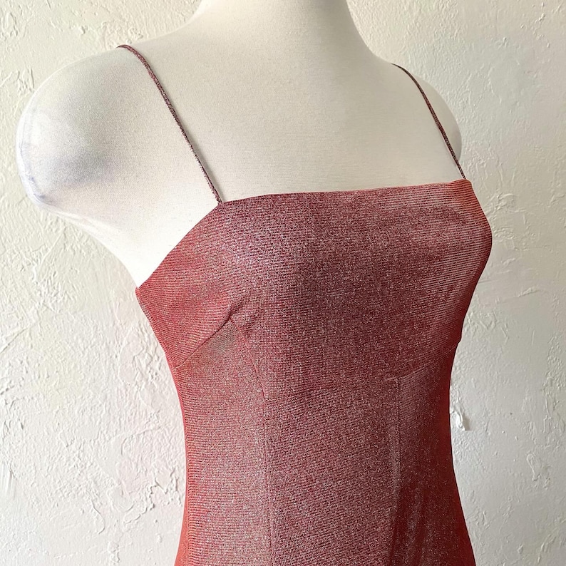 Puede incluir: Vestido rojo-marr&oacute;n brillante con finos tirantes espagueti y escote cuadrado. El vestido se exhibe en un maniqu&iacute;, mostrando su silueta ajustada. La tela tiene una apariencia sutil y texturizada.