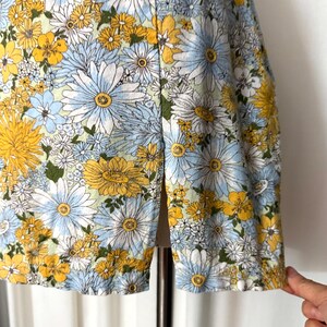May include: A floral print garment featuring a vibrant pattern of yellow, white, and light blue flowers. The fabric is densely covered with various daisy-like blooms, creating a cheerful and summery aesthetic. The garment's hemline is visible, showcasing the detailed floral design.