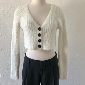 Peut inclure: Un cardigan blanc en fourrure, court, &agrave; col en V et fermeture boutonn&eacute;e. Le cardigan a des manches longues et une texture c&ocirc;tel&eacute;e.