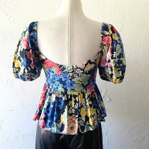 Puede incluir: Una blusa de estampado floral en blanco y negro con un dobladillo con volantes y mangas abullonadas cortas. La blusa tiene un escote cuadrado y un corpi&ntilde;o ajustado.