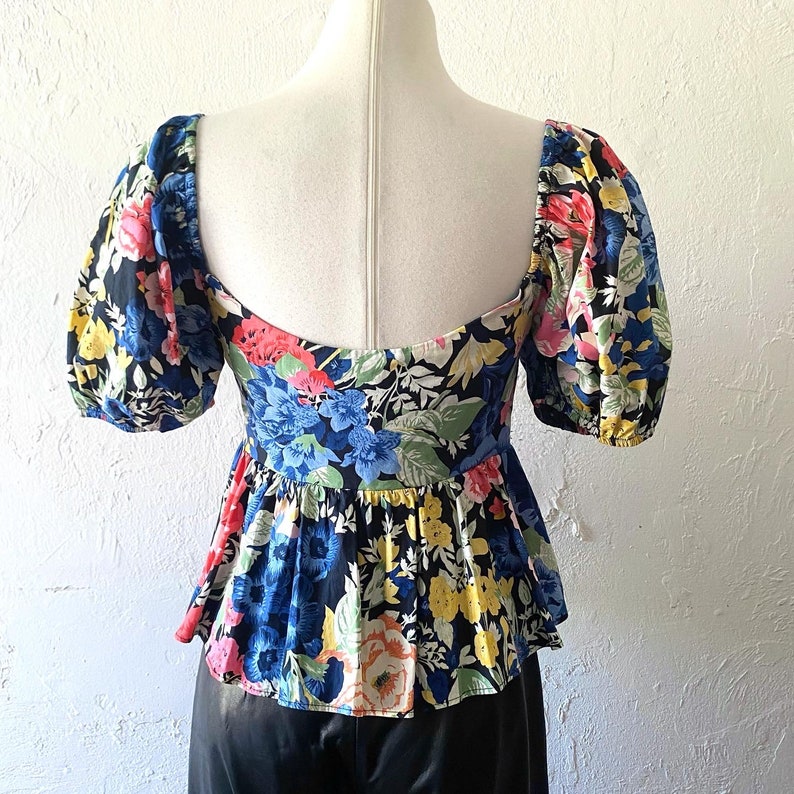 Puede incluir: Una blusa de estampado floral en blanco y negro con un dobladillo con volantes y mangas abullonadas cortas. La blusa tiene un escote cuadrado y un corpi&ntilde;o ajustado.