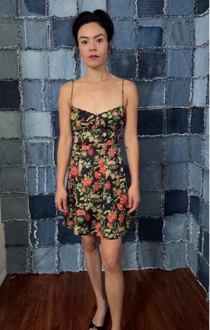 May include: A black floral print dress with spaghetti straps. The dress features a fitted bodice and a flared skirt, adorned with red, yellow, and green floral patterns. The background is a patchwork of denim squares.