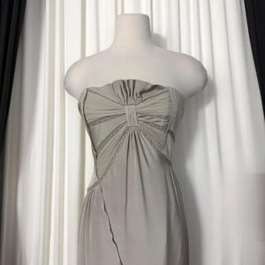 May include: A strapless sage green dress featuring a pleated bodice with a central vertical panel and decorative lace trim accents along the seams. The garment has a fitted silhouette that flows downward from the waist with a smooth fabric finish.