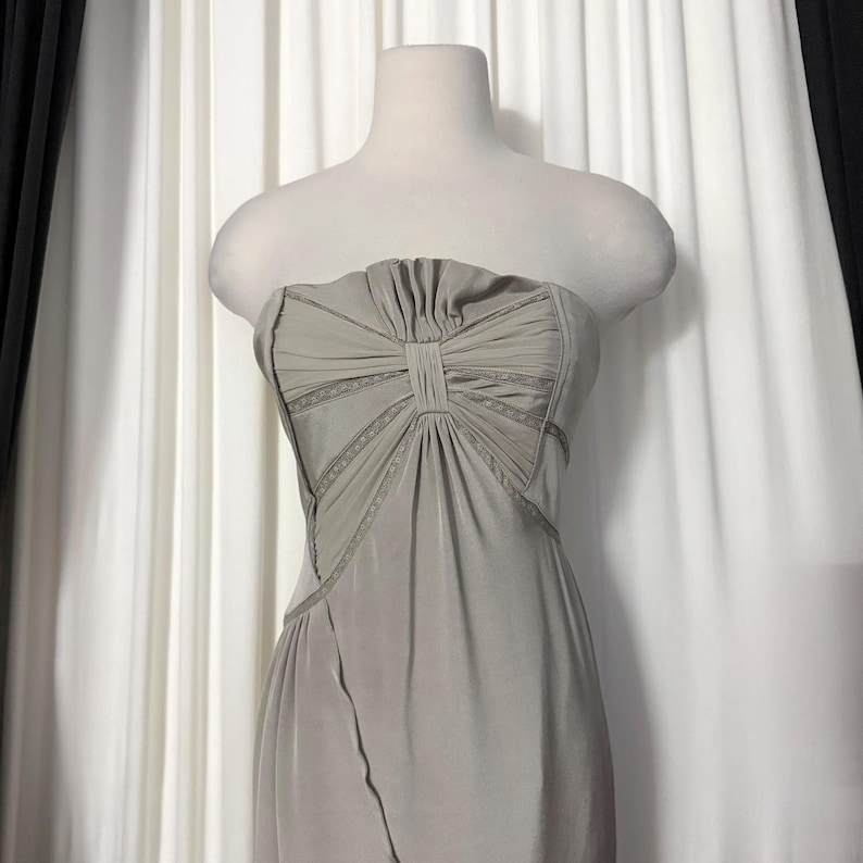 May include: A strapless sage green dress featuring a pleated bodice with a central vertical panel and decorative lace trim accents along the seams. The garment has a fitted silhouette that flows downward from the waist with a smooth fabric finish.