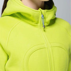 May include: A vibrant lime green hooded sweatshirt with a full zip. The sweatshirt has a high collar, a hood, and a light blue zip pull. The design includes curved seams and a rounded detail on the front.
