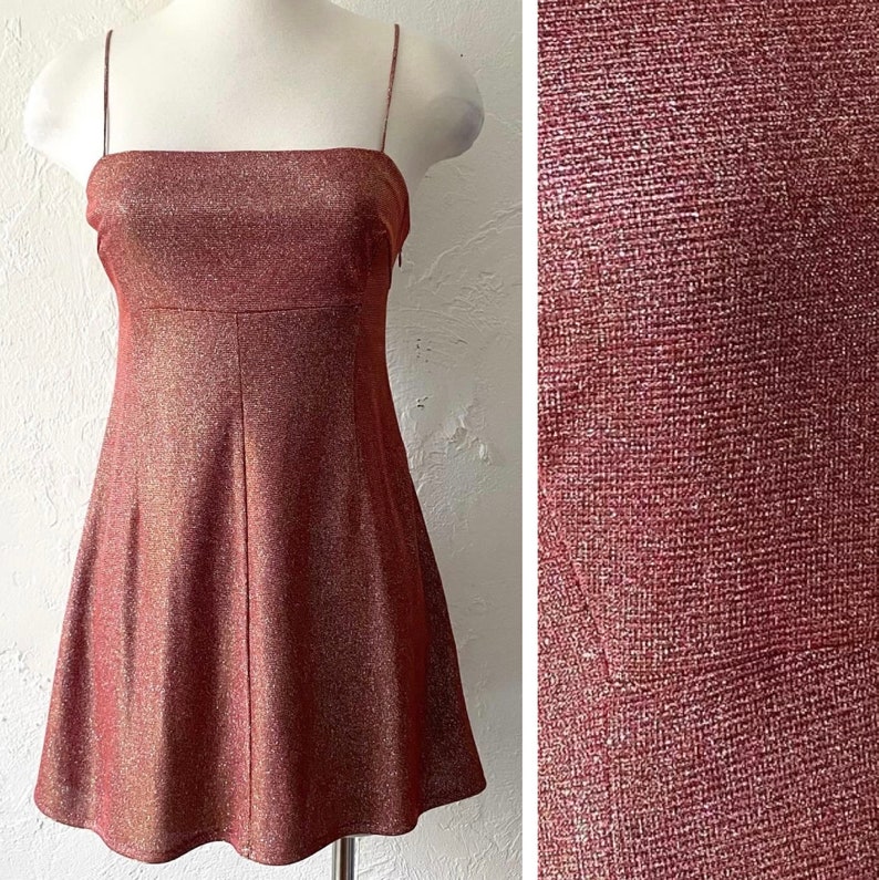 Puede incluir: Un vestido hasta la rodilla con escote cuadrado y finos tirantes espagueti. El vestido es de color marr&oacute;n rojizo con un aspecto brillante y texturizado. El vestido se exhibe en un maniqu&iacute;.