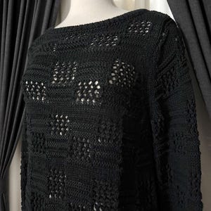 May include: A black knit sweater with a boat neck and long sleeves. The sweater features a textured pattern with openwork details, creating a semi-sheer effect. The garment is displayed on a mannequin against a neutral background.