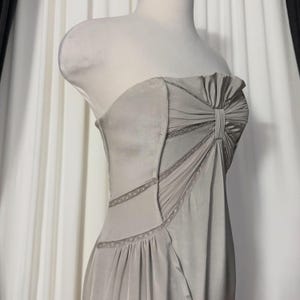 May include: A strapless light gray dress featuring a gathered bodice with a central vertical pleat detail. The garment includes diagonal panels accented with delicate lace trim along the seams, creating a structured yet fluid silhouette.